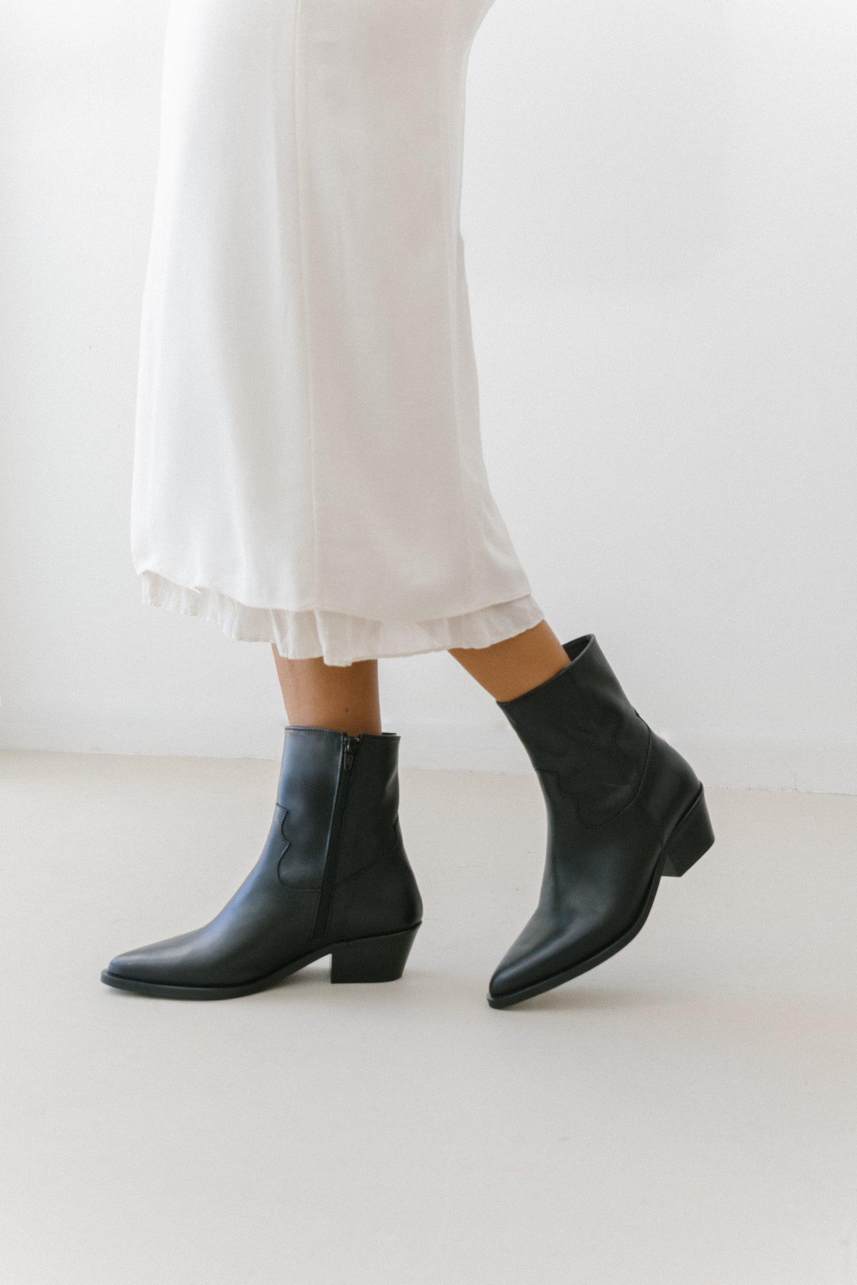Botas texanas pretas em pele, usadas com vestido comprido branco, com o fecho de correr lateral à vista.