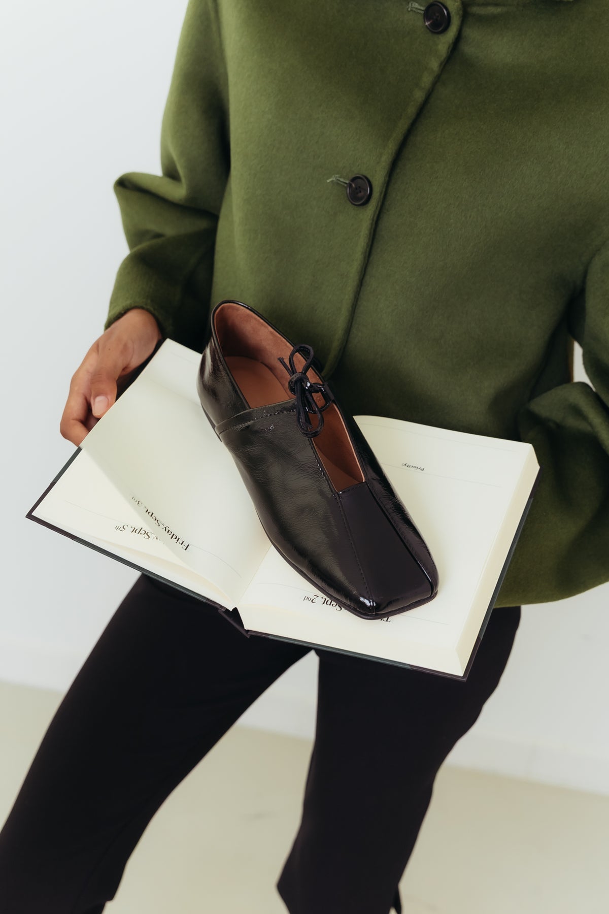 Sapatos rasos pretos em pele envernizada da Zilian, numa imagem de campanha com casaco verde.